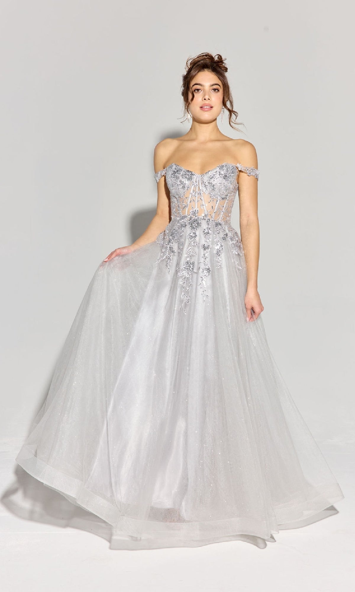 A woman stands against a plain light background in the Eureka EK128 Off-Shoulder Long Prom Ball Gown, featuring a corset-style bodice, embroidered details, sheer accents, and a flowing lace tulle skirt in shimmering silver.